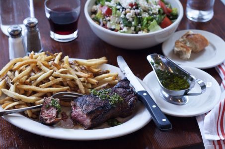 Steak and Fries with chimichurri sauce next to a glass of red wine and salad